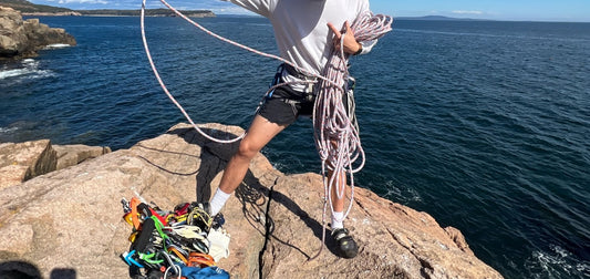 Man coiling rope while standing on a cliffside and wearing socks with climbing shoes.